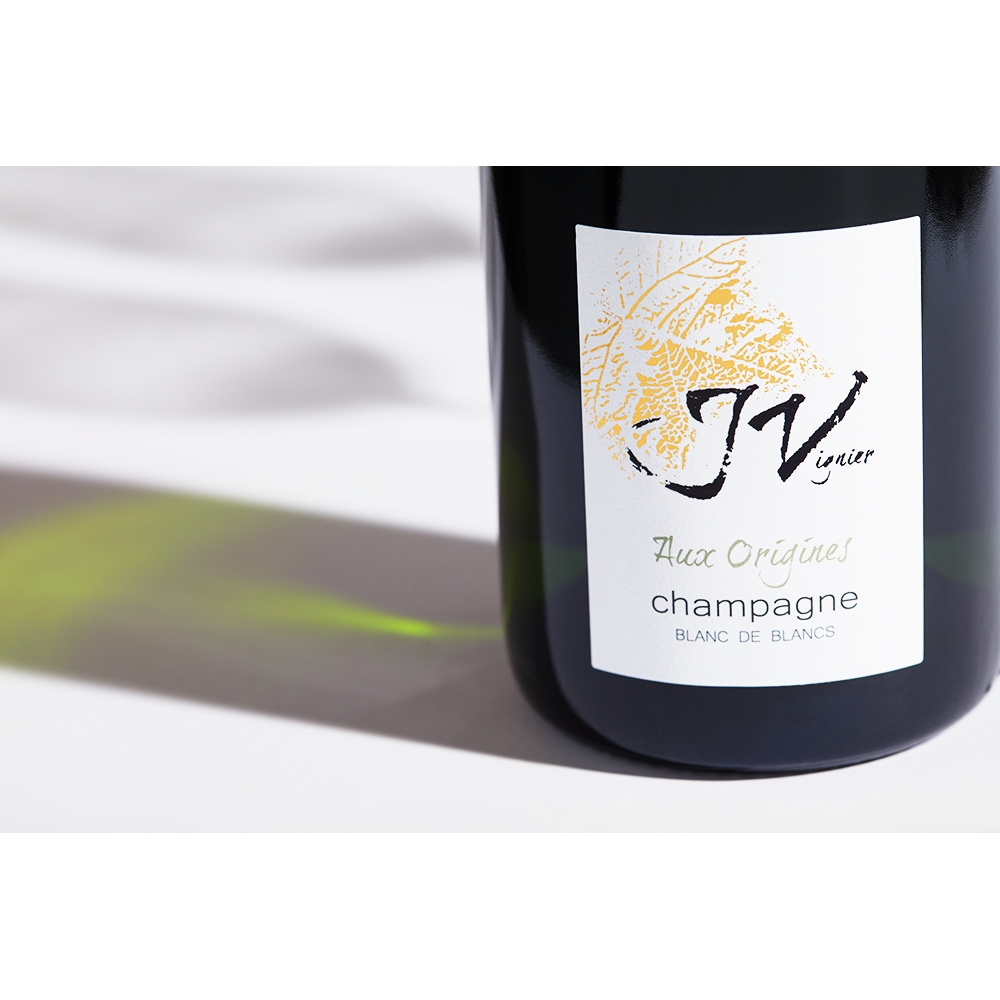 Close up label on a bottle of J Vignier Aux Origines Blanc de Blancs NV on a white table with shadows of the bottle and 2 champagne glasses