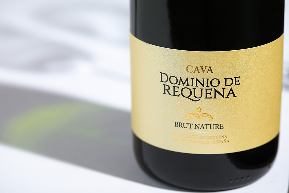 Close up label on a bottle of Pago de Tharsys Dominio Requena Cava Brut Nature Utiel Requena on a white table with shadows of the bottle and 2 champagne glasses