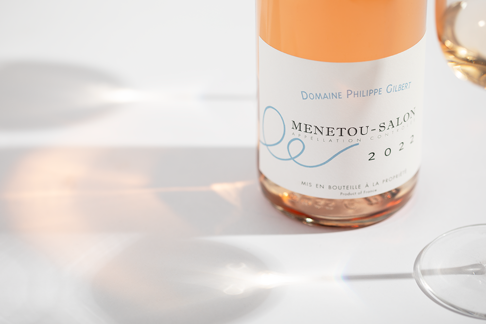 Close up of a bottle of Philippe Gilbert Menetou Salon Rosé 2022 in sunlight on a white background. A glass of rose is also partly seen on the right .