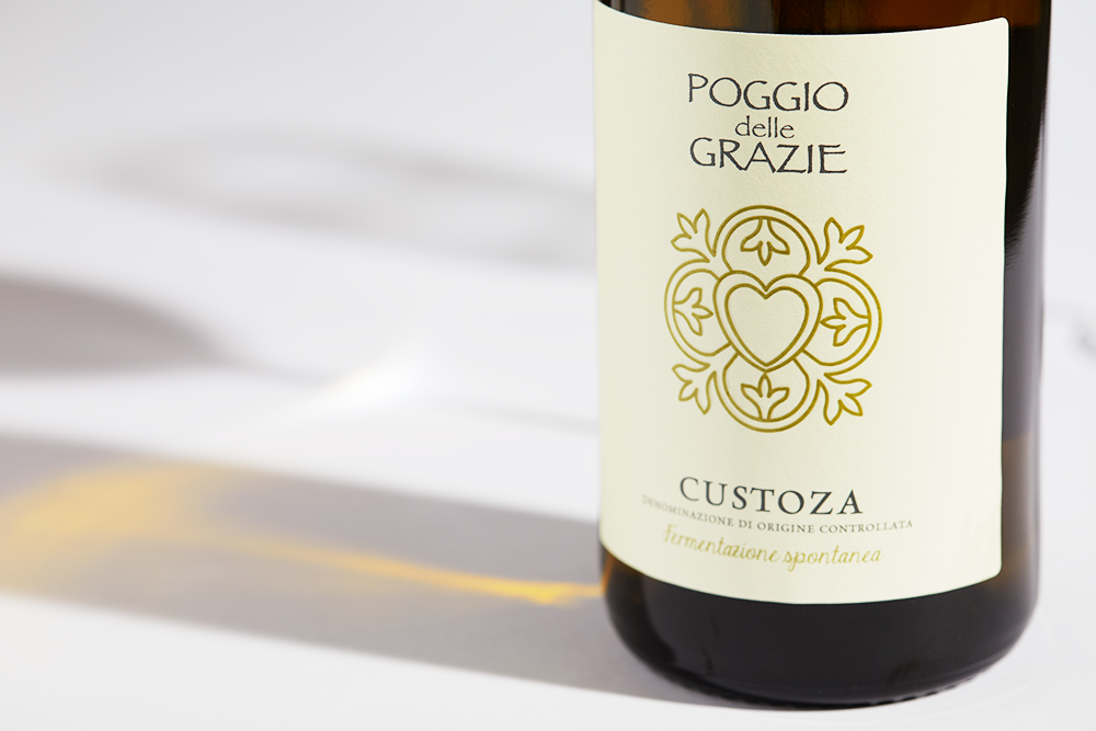 Close up label on a bottle of Poggio delle Grazie Custoza 2022 on a white table with shadows of the bottle and 2 wine glasses