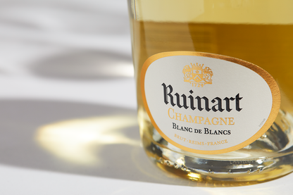 Close up label on a bottle of Ruinart Blanc de Blancs on a white table with shadows of the bottle and 2 champagne glasses