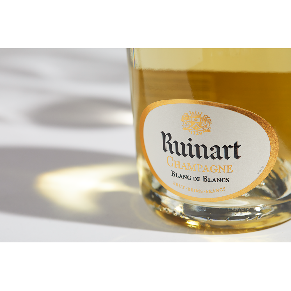 Close up label on a bottle of Ruinart Blanc de Blancs on a white table with shadows of the bottle and 2 champagne glasses