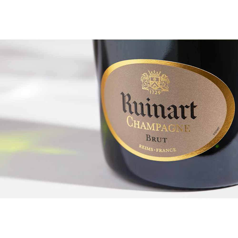 Close up label on a bottle of Ruinart Brut NV on a white table with shadows of the bottle and 2 champagne glasses