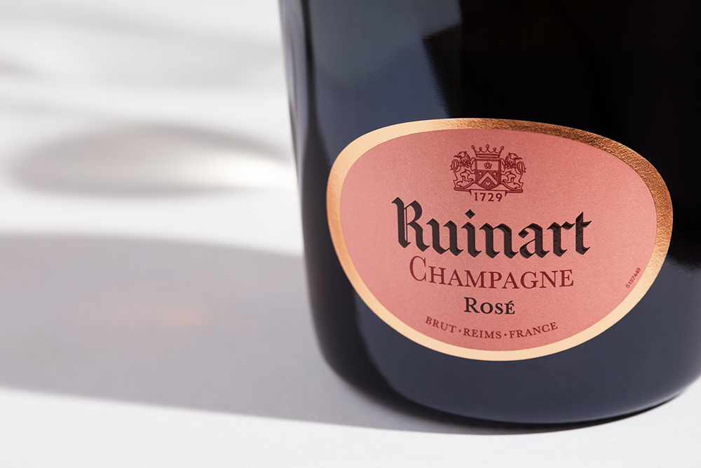 Close up label on a bottle of Ruinart Rosé on a white table with shadows of the bottle and 2 champagne glasses