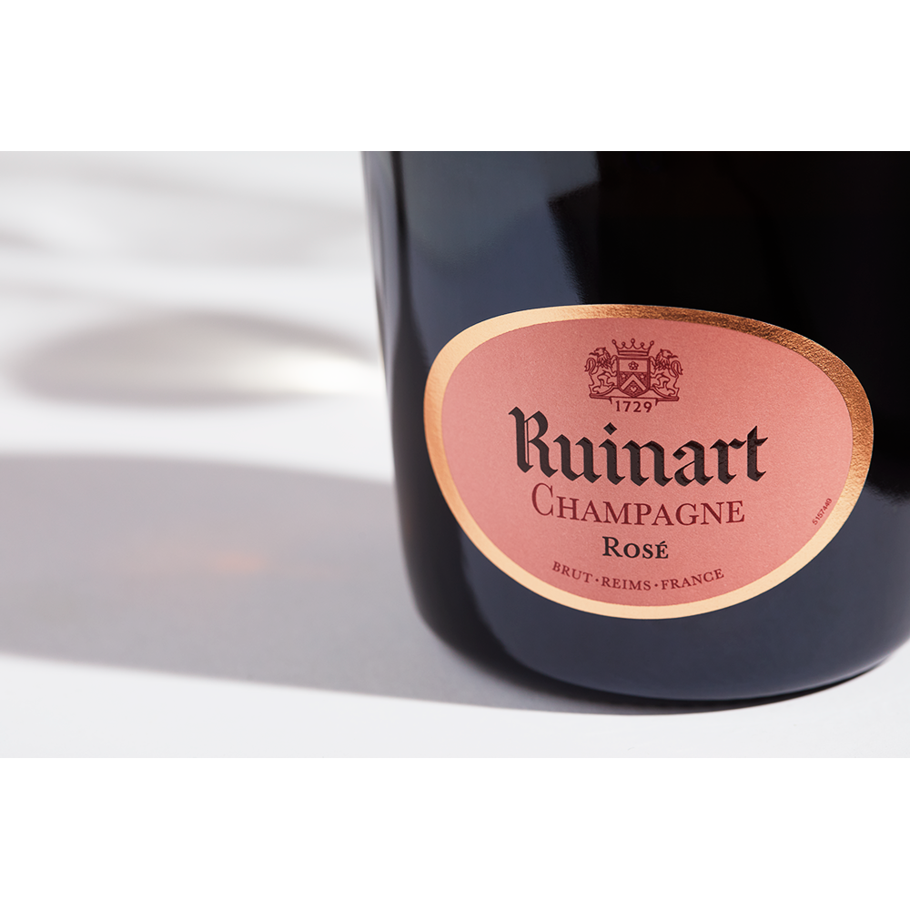 Close up label on a bottle of Ruinart Rosé on a white table with shadows of the bottle and 2 champagne glasses